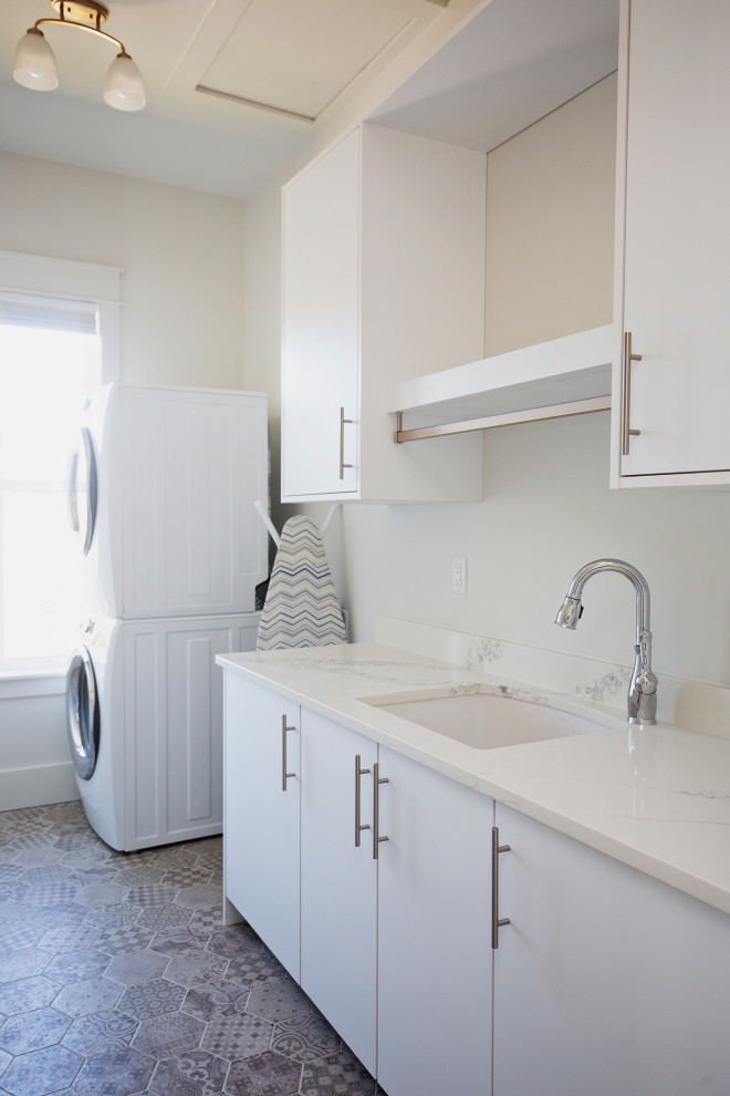 Photo of a mid-sized contemporary single-wall dedicated laundry room in Miami with a drop-in sink, flat-panel cabinets, white cabinets, quartz benchtops, white splashback, engineered quartz splashback, white walls, ceramic floors, a stacked washer and dryer, grey floor and white benchtop.