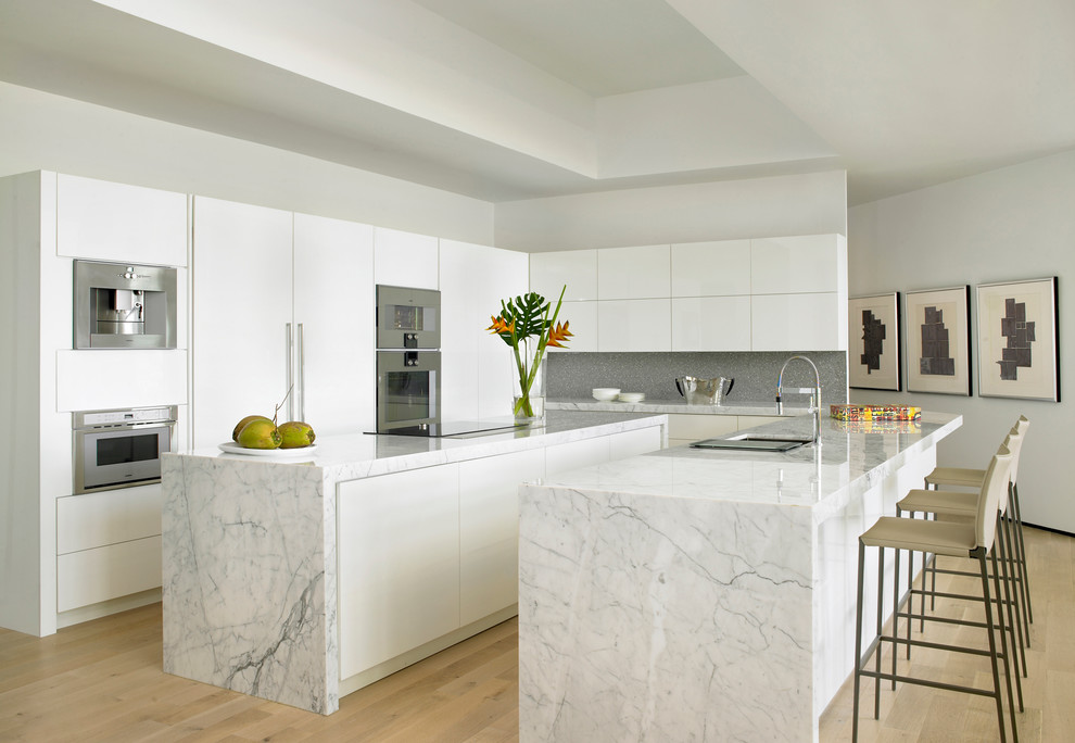 Photo of a large contemporary galley kitchen in Miami with an undermount sink, flat-panel cabinets, white cabinets, grey splashback, stainless steel appliances, light hardwood floors, multiple islands, marble benchtops and beige floor.