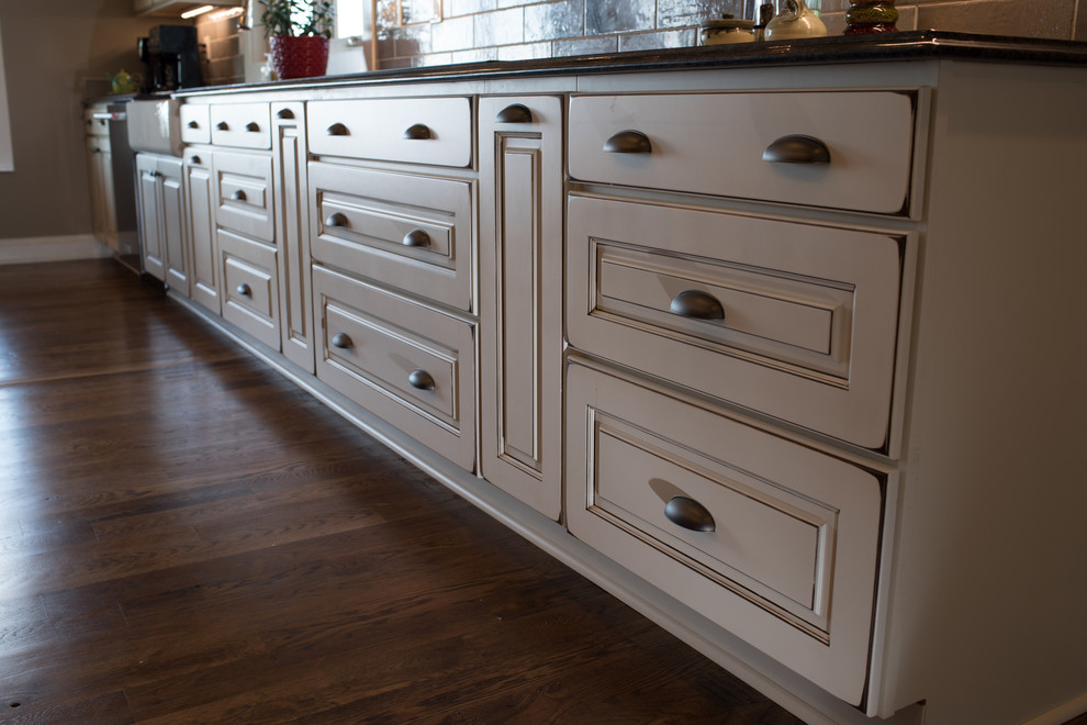 Eat-in kitchen - large rustic l-shaped dark wood floor and brown floor eat-in kitchen idea with a farmhouse sink, raised-panel cabinets, granite countertops, gray backsplash, ceramic backsplash, black appliances, an island and white cabinets