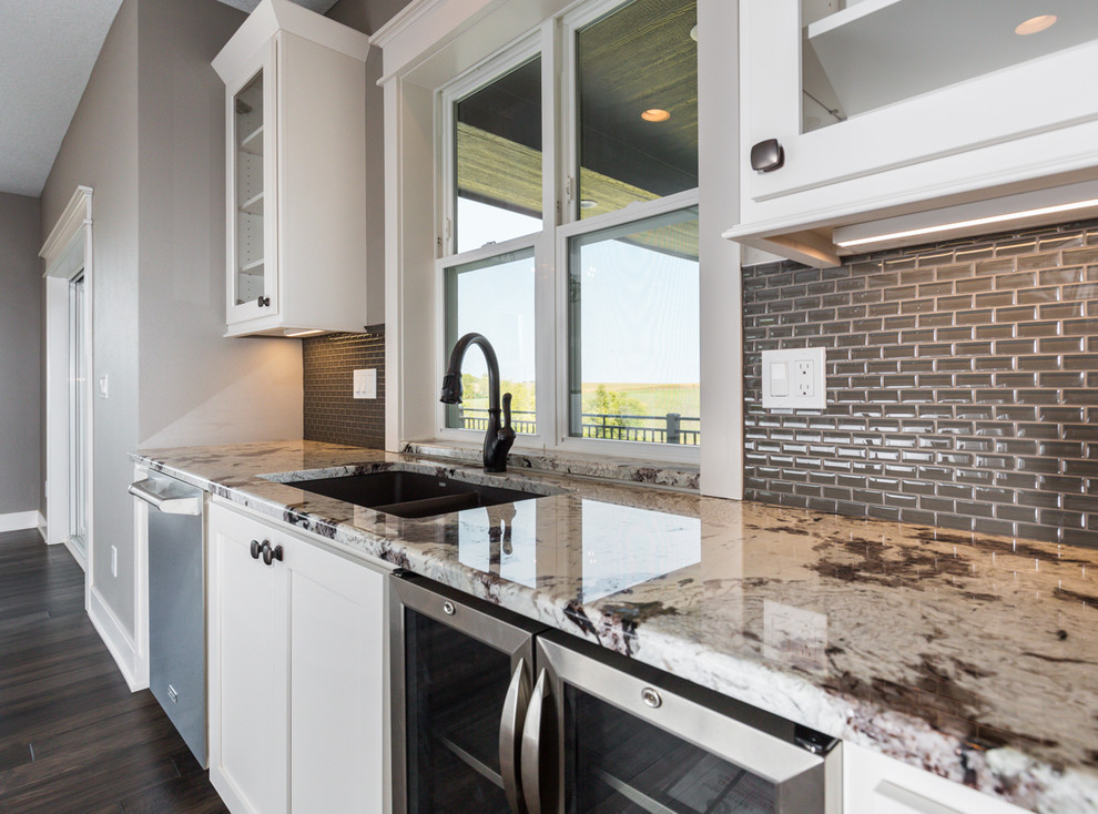 Transitional l-shaped dark wood floor and brown floor kitchen photo with an undermount sink, shaker cabinets, white cabinets, granite countertops, gray backsplash, glass sheet backsplash, stainless steel appliances, an island and white countertops
