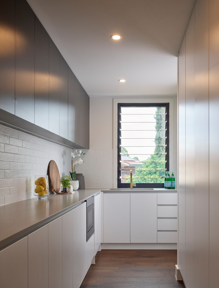 Inspiration for a mid-sized contemporary l-shaped kitchen in Brisbane with an undermount sink, flat-panel cabinets, white cabinets, white splashback, ceramic splashback, panelled appliances, medium hardwood floors, no island, brown floor and grey benchtop.