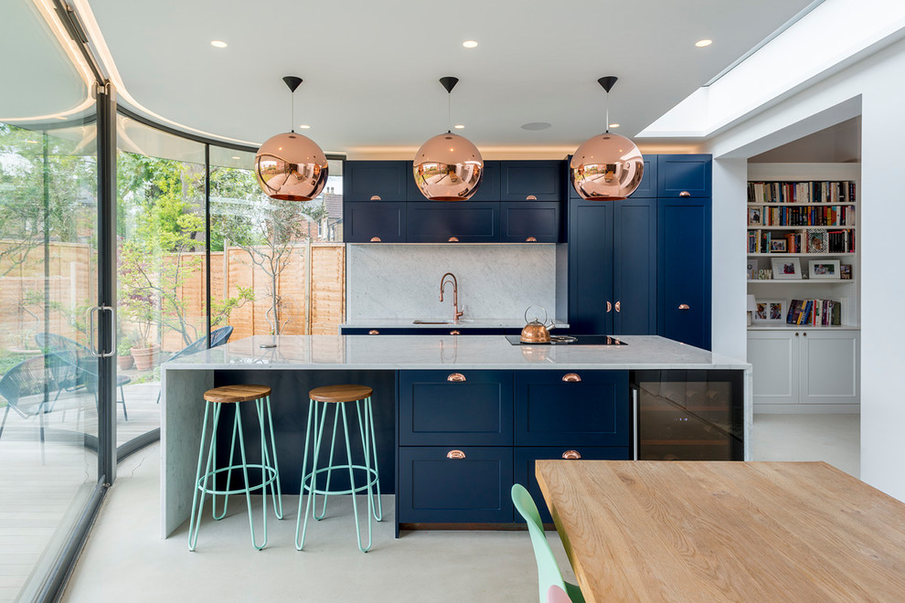 This is an example of a mid-sized contemporary galley open plan kitchen in London with a drop-in sink, shaker cabinets, blue cabinets, marble benchtops, marble splashback, black appliances, concrete floors, with island, grey floor and grey splashback.