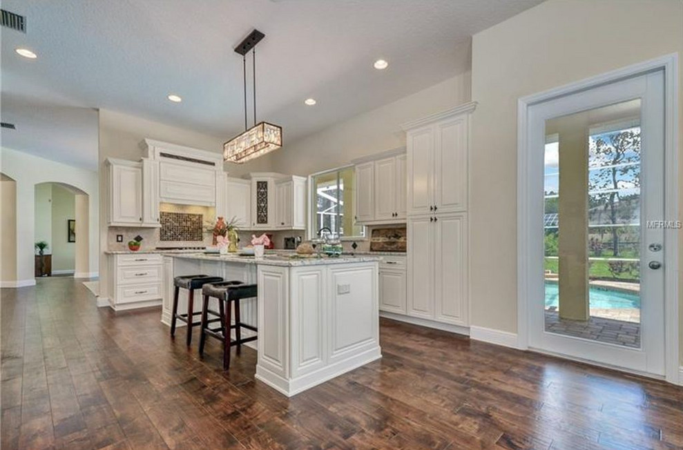 Inspiration for a large transitional l-shaped eat-in kitchen in Tampa with an undermount sink, raised-panel cabinets, beige cabinets, granite benchtops, beige splashback, travertine splashback, stainless steel appliances, dark hardwood floors, with island, brown floor and white benchtop.