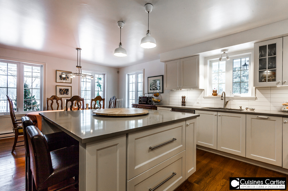 Eat-in kitchen - large transitional medium tone wood floor eat-in kitchen idea in Montreal with a double-bowl sink, shaker cabinets, white cabinets, quartzite countertops, white backsplash, ceramic backsplash, stainless steel appliances and an island
