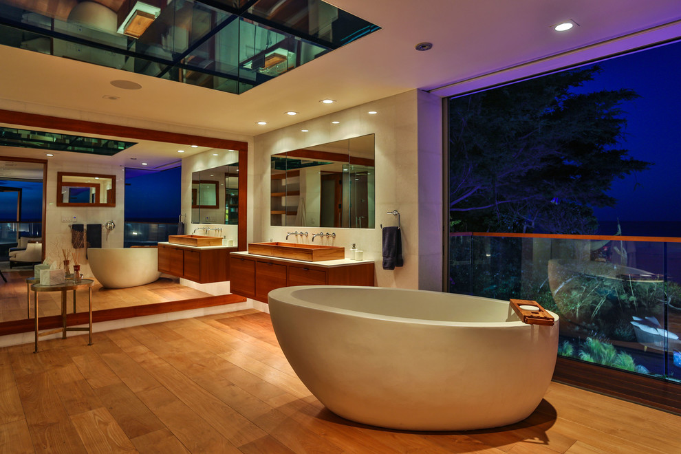 Contemporary master bathroom in Los Angeles with flat-panel cabinets, brown cabinets, a freestanding tub, white tile, ceramic tile, white walls, light hardwood floors, a trough sink, marble benchtops, brown floor and white benchtops.