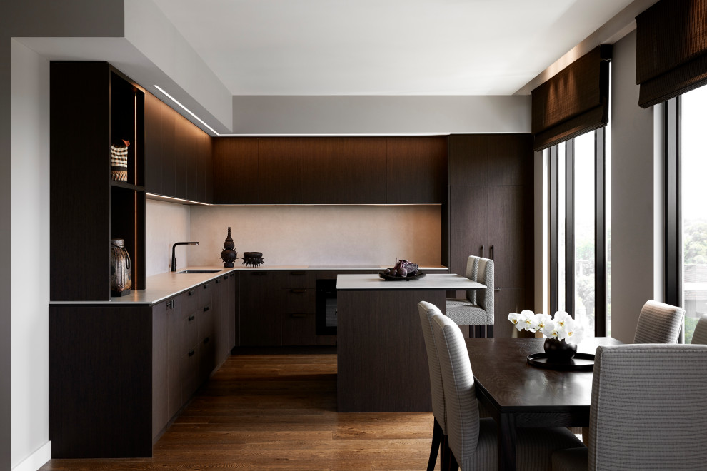 This is an example of a large l-shaped open plan kitchen in Melbourne with an undermount sink, flat-panel cabinets, dark wood cabinets, tile benchtops, beige splashback, ceramic splashback, black appliances, medium hardwood floors, with island, brown floor and beige benchtop.