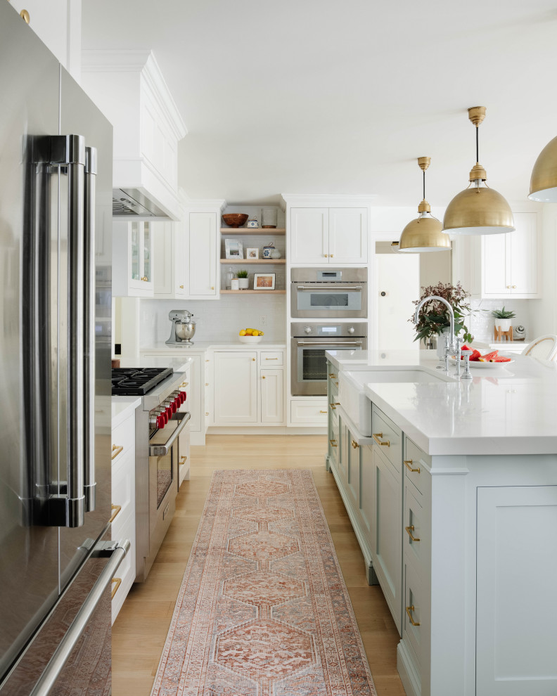 Inspiration for a large traditional l-shaped kitchen/diner in Boston with a belfast sink, recessed-panel cabinets, white cabinets, engineered stone countertops, white splashback, ceramic splashback, stainless steel appliances, light hardwood flooring, an island and white worktops.