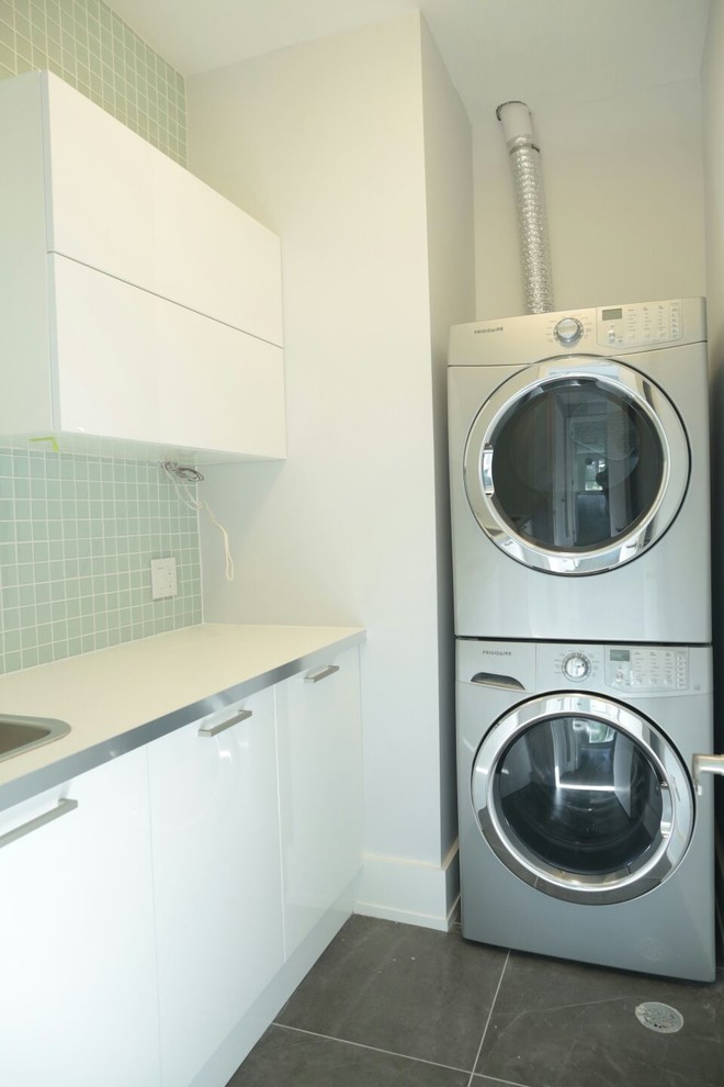 Photo of a mid-sized contemporary l-shaped dedicated laundry room in Toronto with a drop-in sink, flat-panel cabinets, white cabinets, solid surface benchtops, white walls, ceramic floors, a stacked washer and dryer and grey floor.