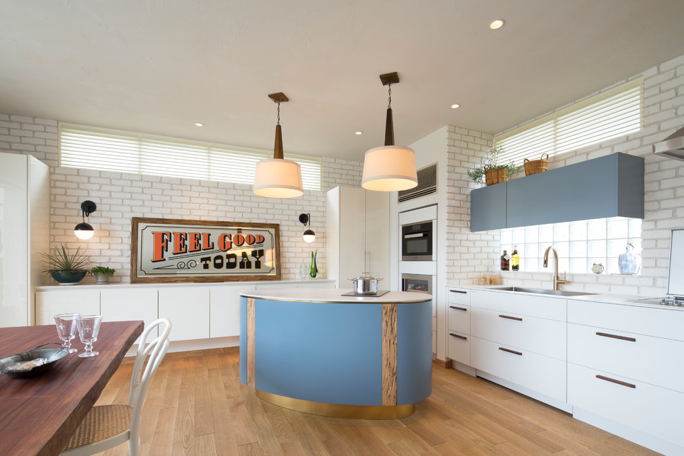 Photo of a beach style single-wall open plan kitchen with an undermount sink, flat-panel cabinets, blue cabinets, white splashback, stainless steel appliances, light hardwood floors, with island and beige floor.