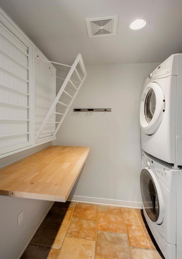 Inspiration for a mid-sized traditional u-shaped dedicated laundry room in Dallas with wood benchtops, grey walls, terra-cotta floors, a stacked washer and dryer, orange floor and brown benchtop.