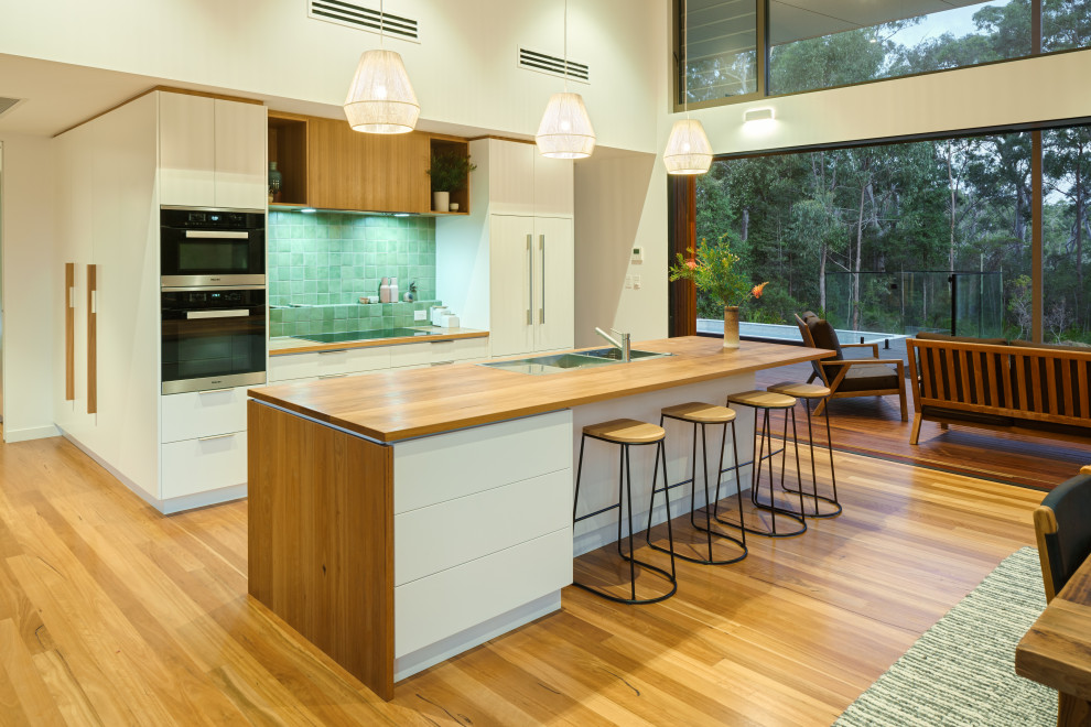 Inspiration for a contemporary galley kitchen in Sunshine Coast with a double-bowl sink, flat-panel cabinets, white cabinets, wood benchtops, green splashback, black appliances, medium hardwood floors, with island, brown floor and brown benchtop.