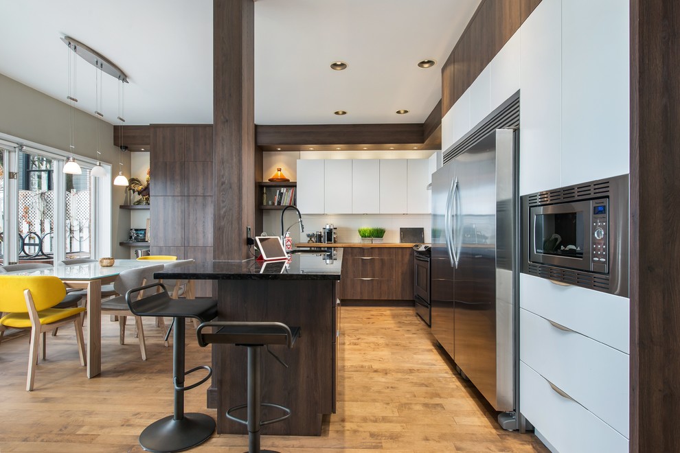 This is an example of a mid-sized contemporary eat-in kitchen in Montreal with an undermount sink, flat-panel cabinets, stainless steel appliances, with island, white cabinets, white splashback, medium hardwood floors and black benchtop.