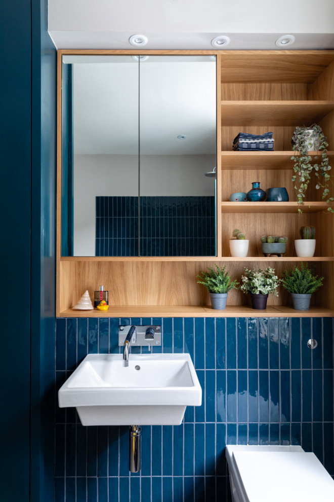 This is an example of a mid-sized transitional kids bathroom in Sussex with flat-panel cabinets, blue cabinets, a drop-in tub, an open shower, a wall-mount toilet, blue tile, ceramic tile, grey walls, porcelain floors, a wall-mount sink, wood benchtops, grey floor, a hinged shower door, blue benchtops, a single vanity and a built-in vanity.