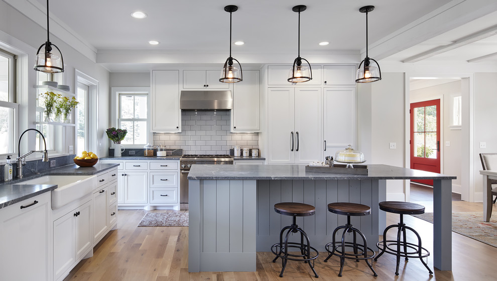 Photo of a mid-sized country l-shaped eat-in kitchen in Minneapolis with a farmhouse sink, white cabinets, soapstone benchtops, white splashback, light hardwood floors, with island, stone tile splashback, panelled appliances, brown floor and recessed-panel cabinets.