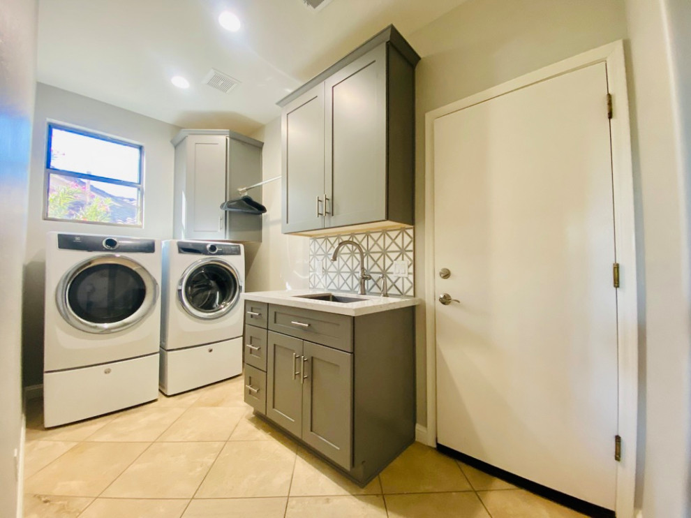 Inredning av en klassisk mellanstor vita l-formad vitt tvättstuga enbart för tvätt, med en undermonterad diskho, skåp i shakerstil, grå skåp, bänkskiva i kvartsit, vitt stänkskydd, stänkskydd i keramik, grå väggar, travertin golv, en tvättmaskin och torktumlare bredvid varandra och beiget golv