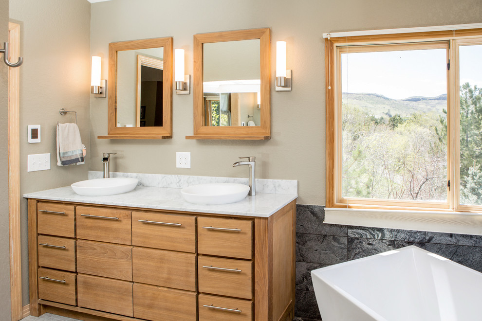 This is an example of a large transitional master bathroom in Denver with flat-panel cabinets, light wood cabinets, a freestanding tub, an alcove shower, a one-piece toilet, black tile, stone tile, grey walls, porcelain floors, a vessel sink, engineered quartz benchtops, grey floor, a hinged shower door and white benchtops.