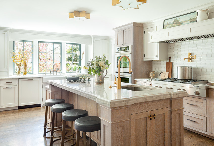 Klassisk inredning av ett vit vitt kök, med en undermonterad diskho, luckor med profilerade fronter, skåp i ljust trä, vitt stänkskydd, stänkskydd i stenkakel, rostfria vitvaror, mellanmörkt trägolv, en köksö, brunt golv och bänkskiva i kvartsit