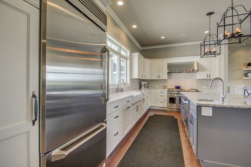 Large transitional l-shaped medium tone wood floor kitchen photo with a farmhouse sink, recessed-panel cabinets, white cabinets, stone tile backsplash, stainless steel appliances and an island