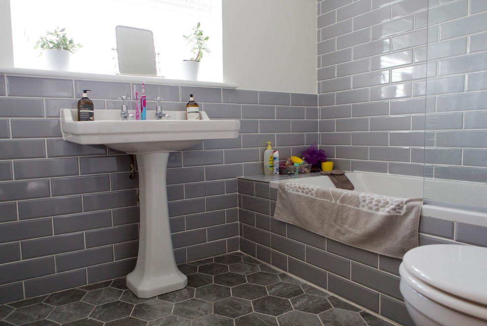 This is an example of a mid-sized traditional kids bathroom in Oxfordshire with recessed-panel cabinets, grey cabinets, a drop-in tub, a shower/bathtub combo, a one-piece toilet, ceramic tile, grey walls, linoleum floors, a pedestal sink, grey floor and a hinged shower door.