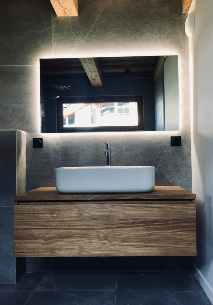 This is an example of a large contemporary master bathroom in Lyon with light wood cabinets, a curbless shower, a wall-mount toilet, gray tile, stone tile, grey walls, ceramic floors, a drop-in sink, wood benchtops, black floor, beige benchtops, a single vanity, a floating vanity and wood.