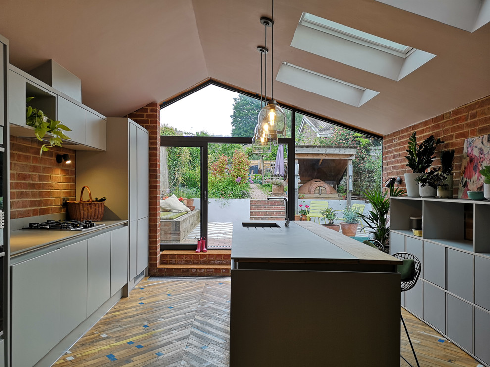 Mid-sized contemporary galley eat-in kitchen in Gloucestershire with an undermount sink, flat-panel cabinets, grey cabinets, tile benchtops, grey splashback, ceramic splashback, panelled appliances, medium hardwood floors, with island, multi-coloured floor and grey benchtop.