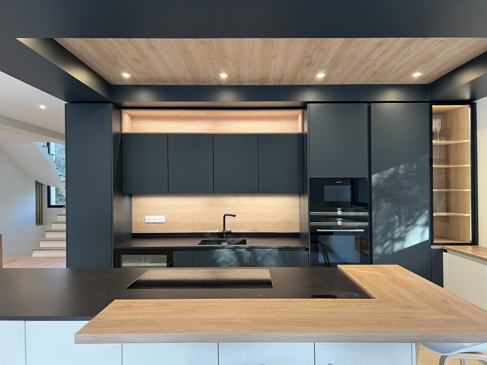 Immagine di una grande cucina contemporanea con ante a filo, ante nere, paraspruzzi beige, paraspruzzi in legno, elettrodomestici neri, pavimento beige, top nero e soffitto ribassato
