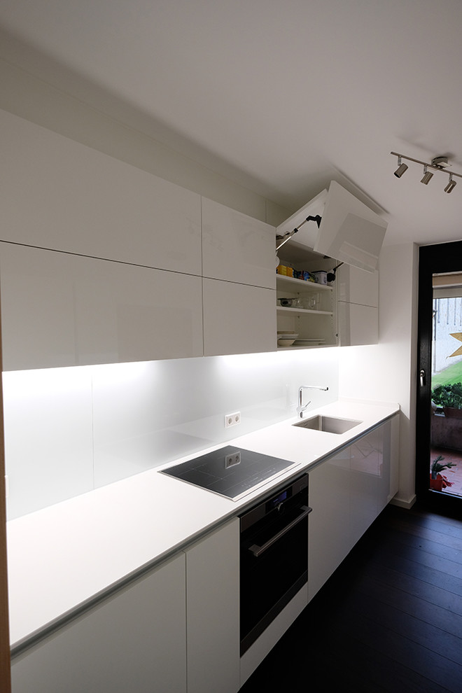 Inspiration for a mid-sized contemporary galley separate kitchen in Other with flat-panel cabinets, white cabinets, quartz benchtops, white benchtop, a drop-in sink, white splashback, glass sheet splashback, black appliances, dark hardwood floors and brown floor.