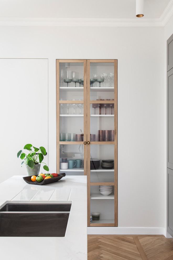 This is an example of a scandinavian galley kitchen in Paris with an integrated sink, grey cabinets, quartzite benchtops, white splashback, panelled appliances, light hardwood floors, with island, white benchtop and glass-front cabinets.