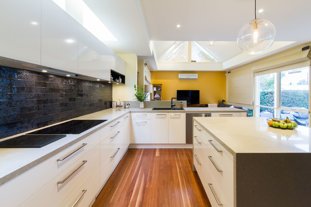 Photo of a large contemporary u-shaped open plan kitchen in Melbourne with flat-panel cabinets, quartz benchtops, black splashback, subway tile splashback, medium hardwood floors, with island and grey benchtop.