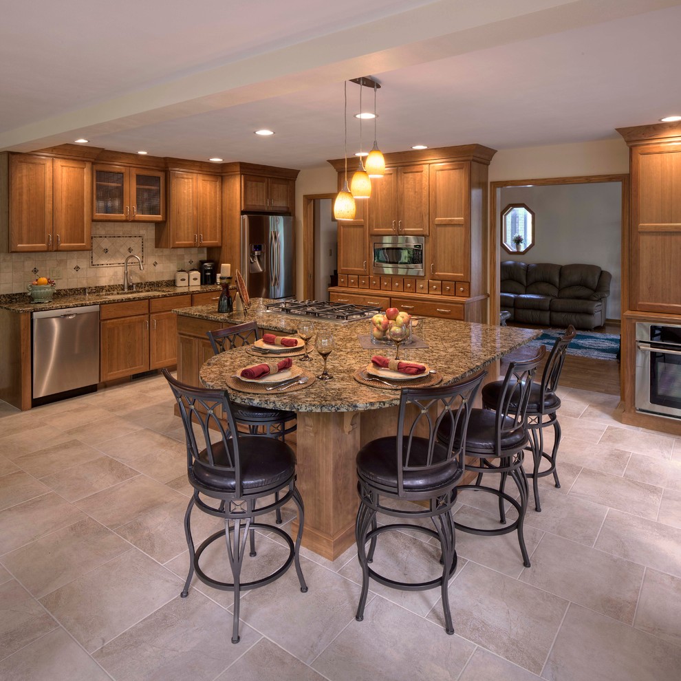 Large traditional l-shaped open plan kitchen in Milwaukee with an undermount sink, recessed-panel cabinets, medium wood cabinets, granite benchtops, beige splashback, ceramic splashback, stainless steel appliances, ceramic floors, with island, beige floor and brown benchtop.