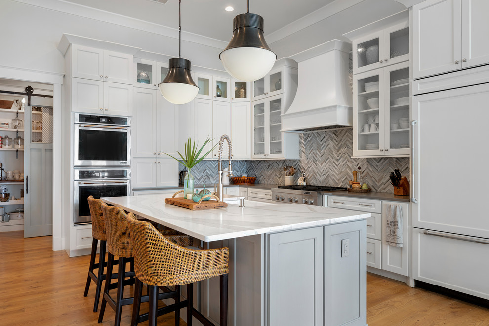Photo of a large beach style u-shaped eat-in kitchen in Charleston with white cabinets, quartzite benchtops, grey splashback, mosaic tile splashback, stainless steel appliances, medium hardwood floors, with island, brown floor and a drop-in sink.