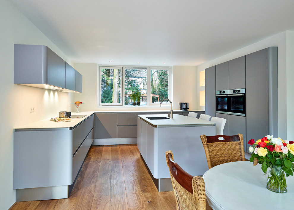Large contemporary u-shaped open plan kitchen in Amsterdam with glass benchtops, medium hardwood floors, with island, a drop-in sink, flat-panel cabinets, grey cabinets and black appliances.