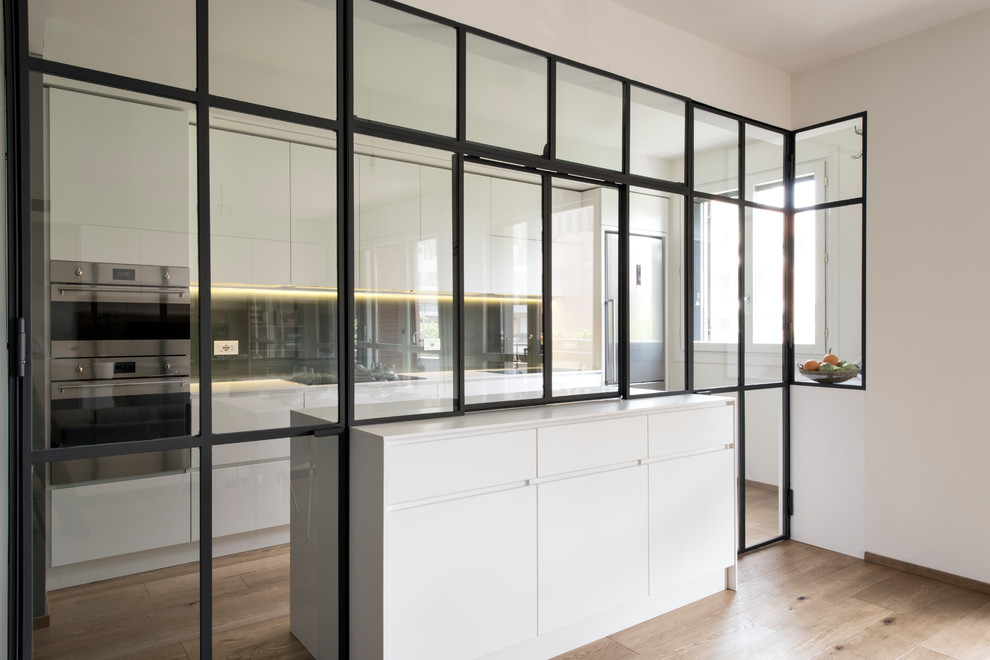 This is an example of a large modern galley separate kitchen in Milan with a drop-in sink, flat-panel cabinets, white cabinets, solid surface benchtops, grey splashback, glass sheet splashback, stainless steel appliances, light hardwood floors and with island.
