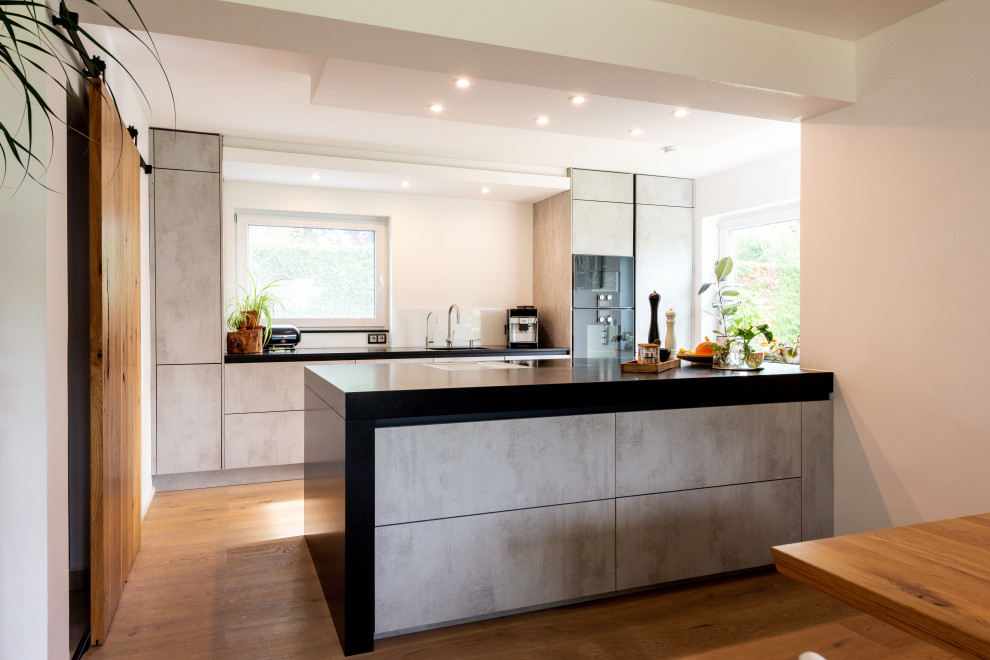 Photo of a contemporary galley kitchen in Munich with flat-panel cabinets, grey cabinets, granite benchtops, black appliances, medium hardwood floors, with island and black benchtop.