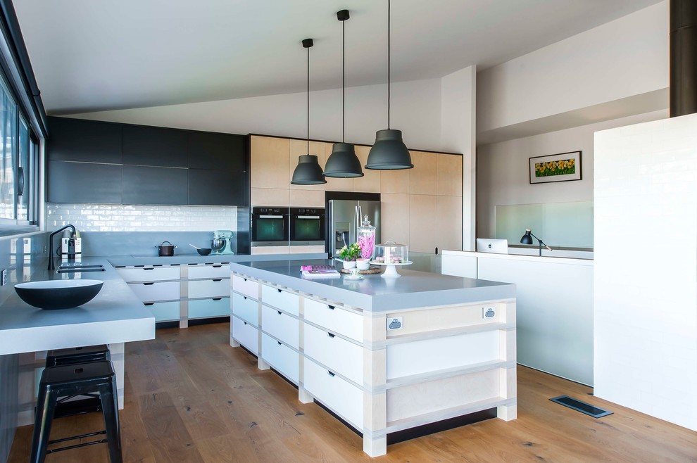 Large contemporary l-shaped eat-in kitchen in Sydney with an undermount sink, flat-panel cabinets, light wood cabinets, solid surface benchtops, grey splashback, porcelain splashback, black appliances, light hardwood floors and with island.