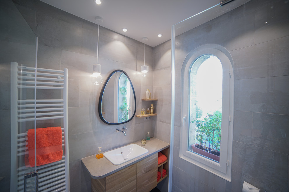 This is an example of a small contemporary 3/4 bathroom in Paris with beaded inset cabinets, light wood cabinets, a wall-mount toilet, gray tile, porcelain tile, grey walls, porcelain floors, an undermount sink, limestone benchtops, grey floor, a hinged shower door and a corner shower.