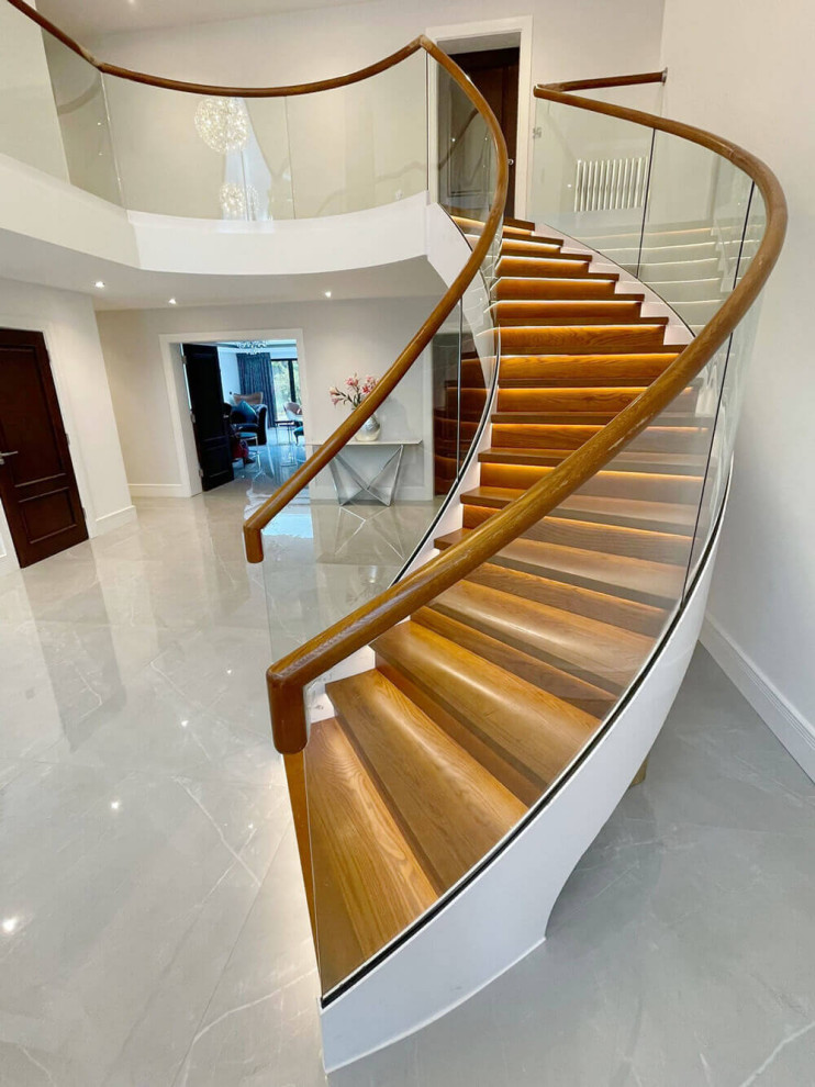 Réalisation d'un grand escalier courbe minimaliste avec des marches en bois, des contremarches en bois, un garde-corps en verre et du lambris.