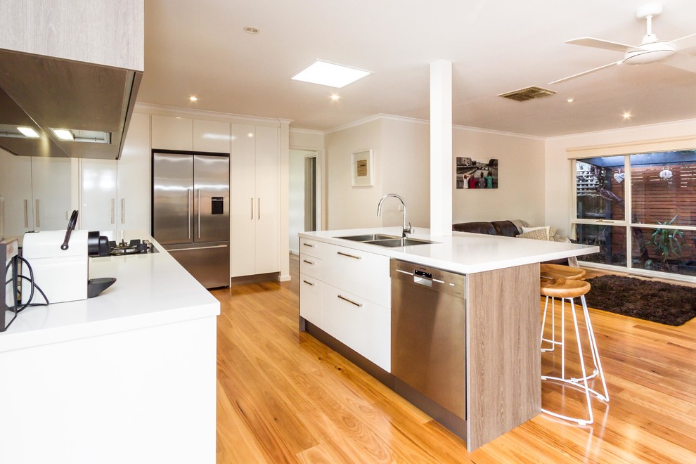 This is an example of a mid-sized contemporary l-shaped kitchen pantry in Melbourne with a drop-in sink, flat-panel cabinets, white cabinets, quartz benchtops, brown splashback, glass sheet splashback, stainless steel appliances, medium hardwood floors, with island and brown floor.
