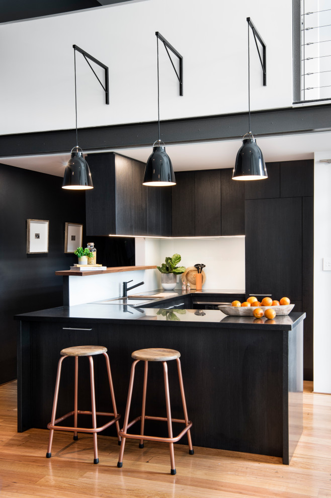 Inspiration for a small industrial u-shaped eat-in kitchen in Sydney with a drop-in sink, white splashback, glass sheet splashback, black appliances, medium hardwood floors, a peninsula and black benchtop.