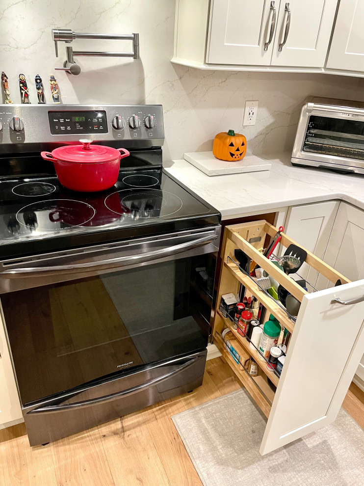 Transitional l-shaped light wood floor and beige floor eat-in kitchen photo with a farmhouse sink, shaker cabinets, white cabinets, quartz countertops, white backsplash, quartz backsplash, black appliances, an island and white countertops