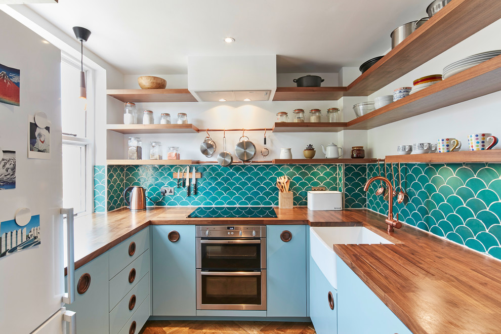 Inspiration for a small midcentury u-shaped separate kitchen in London with a farmhouse sink, flat-panel cabinets, blue cabinets, wood benchtops, stainless steel appliances, medium hardwood floors, no island, brown floor, green splashback and ceramic splashback.