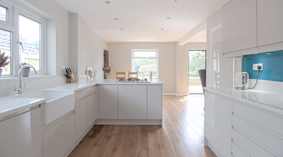 Design ideas for a large modern l-shaped open plan kitchen in Essex with a farmhouse sink, flat-panel cabinets, white cabinets, quartzite benchtops, blue splashback, glass sheet splashback, stainless steel appliances, vinyl floors and a peninsula.