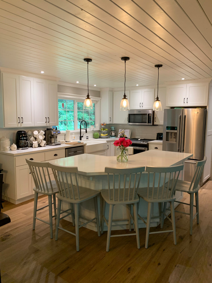 Mid-sized country l-shaped eat-in kitchen in Other with a farmhouse sink, raised-panel cabinets, white cabinets, quartz benchtops, white splashback, subway tile splashback, stainless steel appliances, medium hardwood floors, with island, brown floor and white benchtop.