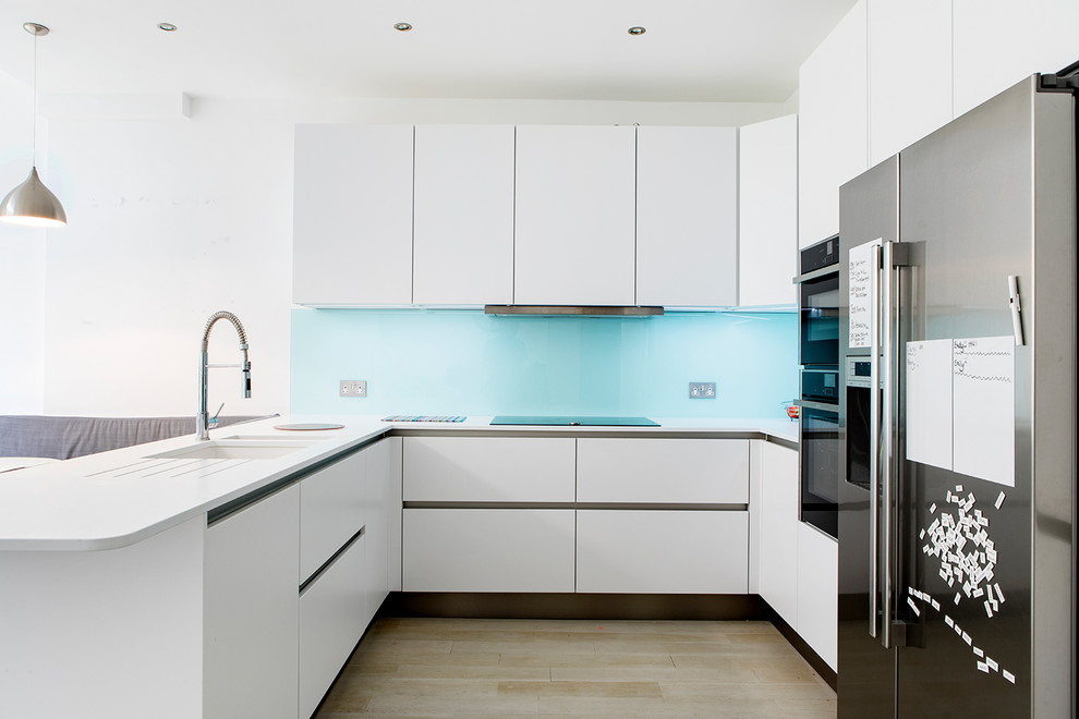 This is an example of a small contemporary u-shaped open plan kitchen in London with blue splashback, no island, a single-bowl sink, flat-panel cabinets, white cabinets, quartzite benchtops, glass sheet splashback and stainless steel appliances.