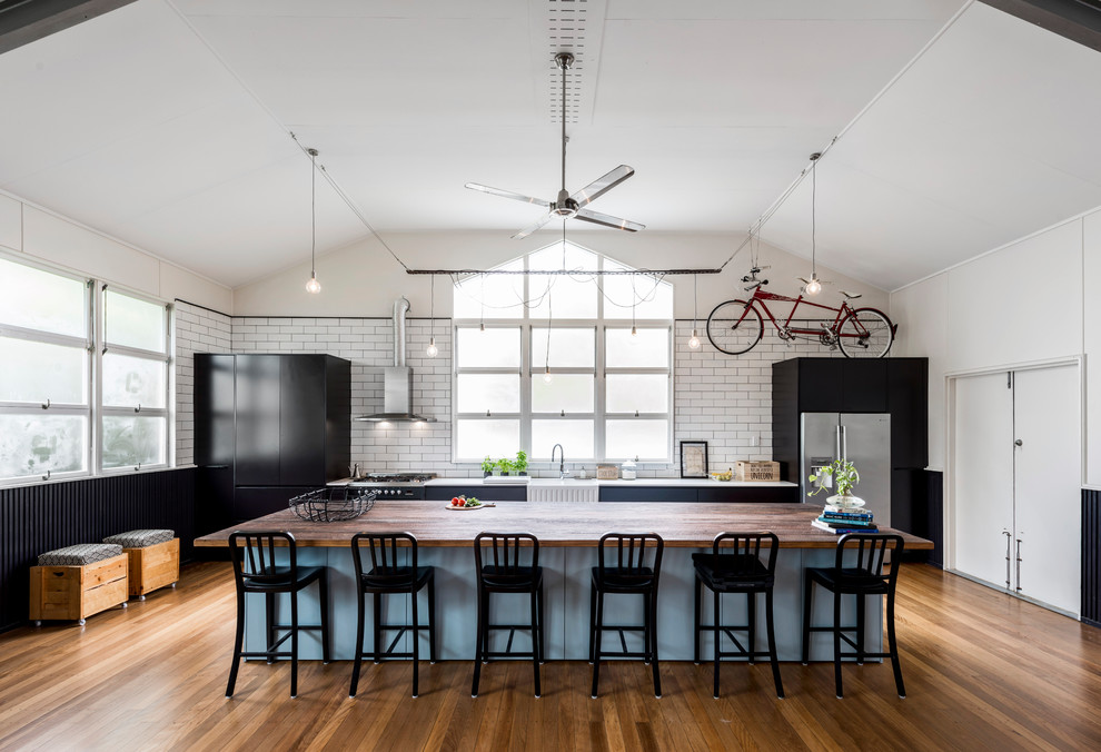 This is an example of a large industrial galley open plan kitchen in Brisbane with a farmhouse sink, flat-panel cabinets, black cabinets, quartz benchtops, white splashback, ceramic splashback, stainless steel appliances, medium hardwood floors and with island.