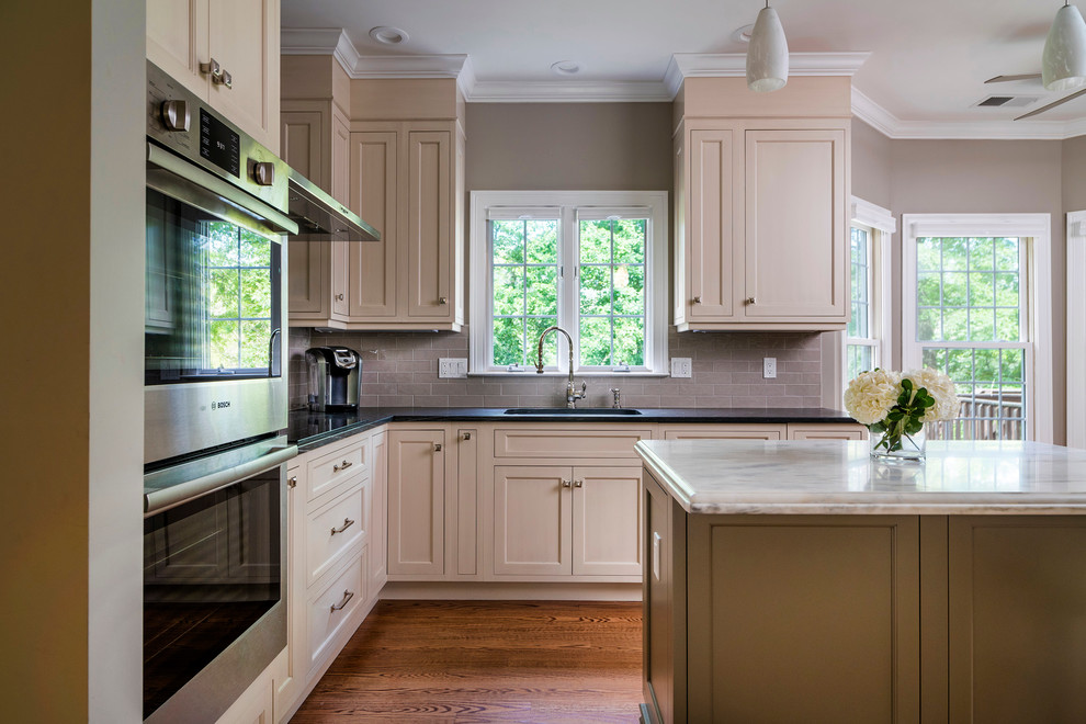 Open concept kitchen - small transitional u-shaped medium tone wood floor and brown floor open concept kitchen idea with an island, an undermount sink, beaded inset cabinets, beige cabinets, gray backsplash, ceramic backsplash, stainless steel appliances and limestone countertops