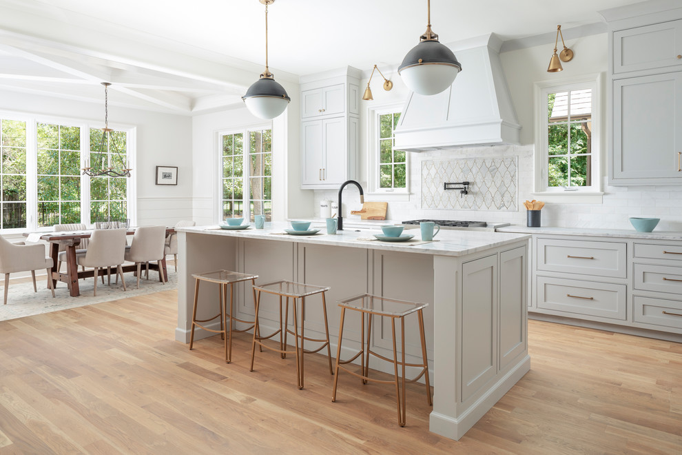 This is an example of a large transitional l-shaped open plan kitchen in Charlotte with a farmhouse sink, grey cabinets, marble benchtops, marble splashback, stainless steel appliances, medium hardwood floors, with island, beaded inset cabinets, white splashback, brown floor and white benchtop.