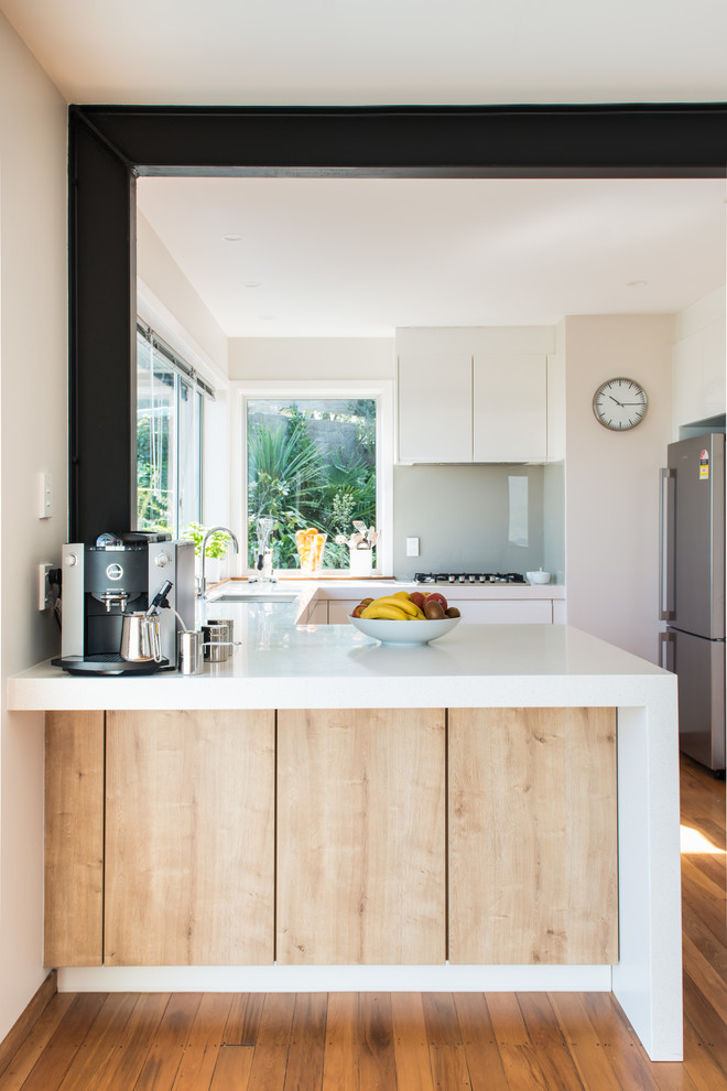 Inspiration for a mid-sized scandinavian u-shaped kitchen pantry in Christchurch with an undermount sink, flat-panel cabinets, white cabinets, solid surface benchtops, glass sheet splashback, white appliances, medium hardwood floors, a peninsula and brown floor.