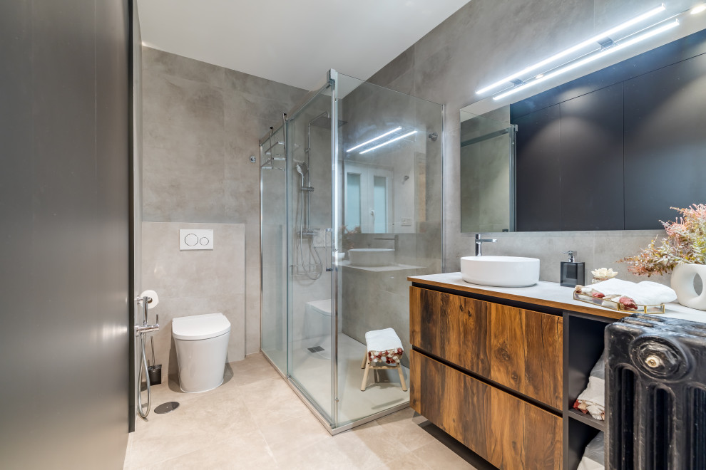 Design ideas for a large urban grey and black ensuite bathroom in Madrid with dark wood cabinets, a corner shower, a wall mounted toilet, grey tiles, grey walls, porcelain flooring, a vessel sink, tiled worktops, beige floors, a sliding door, grey worktops, a single sink, a built in vanity unit and wood walls.