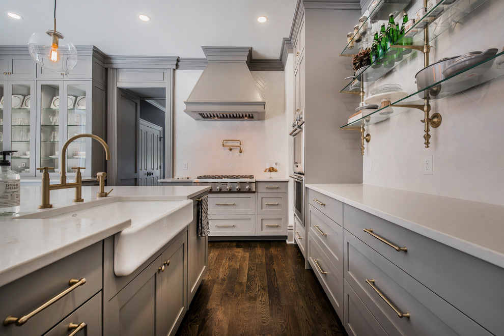Design ideas for a transitional l-shaped kitchen in Columbus with a farmhouse sink, shaker cabinets, grey cabinets, white splashback, stainless steel appliances, dark hardwood floors, with island, brown floor and subway tile splashback.